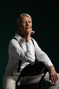 Senior female sitting on black folding chair, posing in studio wearing white shirt and suspenders