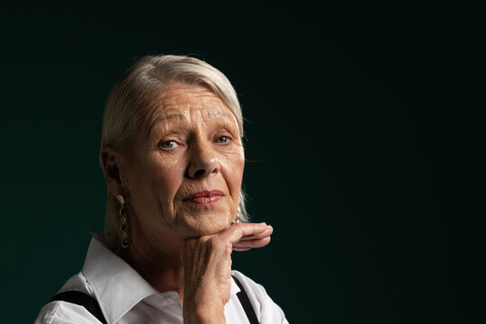 Senior woman posing in studio, resting chin, wearing white shirt, straps, earrings, copy space