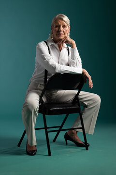 Senior woman sitting backward on black folding chair, posing in teal studio in suspenders and heels