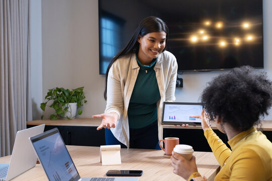 African American female coworkers presenting tablet showing charts at conference table with laptops