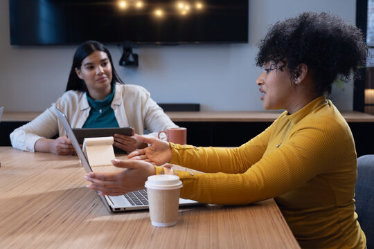 Diverse female colleagues discussing project in modern meeting room, gesturing by laptop and tablet