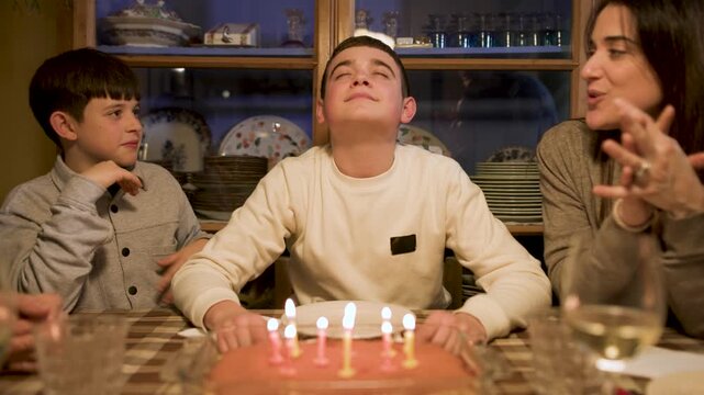 Close-up shot of a teenage boy with closed eyes, making a wish and blowing out his birthday candles as his younger brother and mother watch nearby. Slow motion, video 4K.