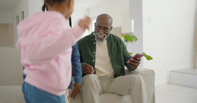 Senior African American man with two children tapping phone encouraging playing and dancing at home