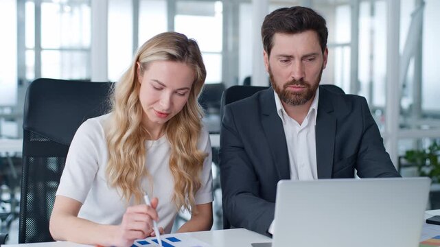 Successful two business people entrepreneurs looking in laptop internet cyberspace talking chatting comparing data in computer and documents diagram graphs Caucasian man talk with woman talk in office