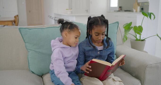 African American sisters sitting on sofa, older opening red book, reading aloud, teaching younger