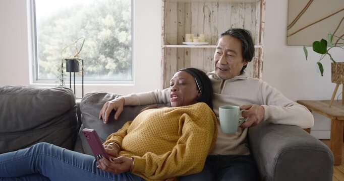 Diverse couple senior-man resting on couch, woman scrolling purple phone, man holding mug, pointing