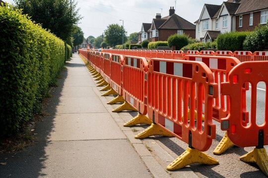 Orange plastic safety barriers line sidewalk near roadwork site. Construction fences separate pedestrian pavement from street in residential area. Suburban houses, green hedge in background. Traffic