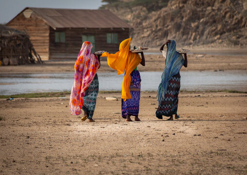 Three women carry trays of shell necklaces on their shoulders while walking past a wooden house, Northern Red Sea, Dahlak, Eritrea
