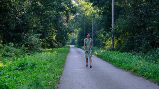 Young woman in green summer dress walks across rustic road . Lush green meadow and forest trees surround narrow dirt path in countryside park.