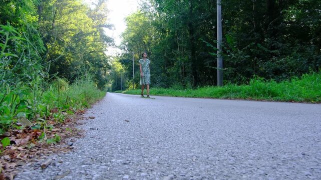 Young woman in green summer dress walks across rustic road . Lush green meadow and forest trees surround narrow dirt path in countryside park.