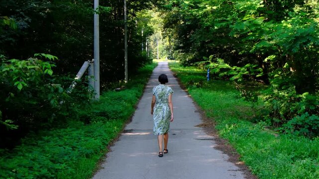 Young woman in green summer dress walks across rustic road . Lush green meadow and forest trees surround narrow dirt path in countryside park.