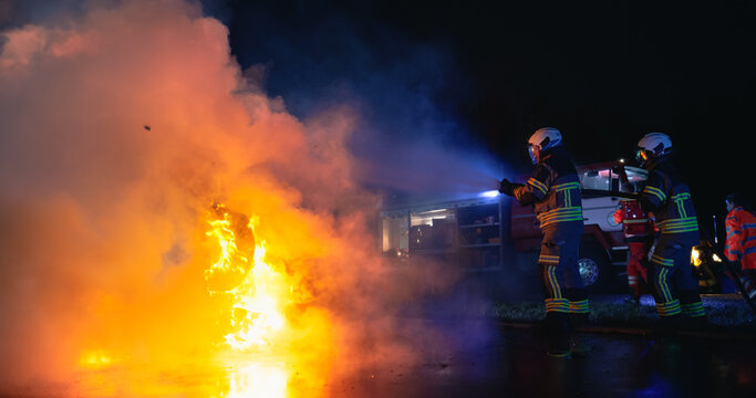 Car Crash: Firefighters in Protective Gear Spray a Water Hose to Extinguish Flames From a Burning Car at a Night, With a Fire Truck and Paramedic Standing by During the Emergency Rescue Operation.