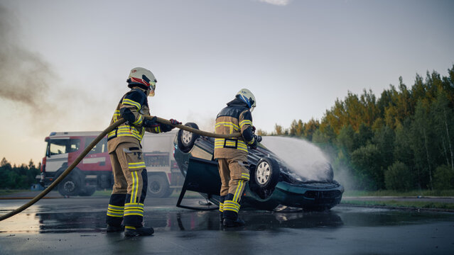 Car Crash: Two Firefighters in White Helmets Spray Water From a Hose Onto an Upside Down Wrecked Car After a Highway Rollover Crash, With a Red Fire Truck and Smoke in the Background at Dusk.