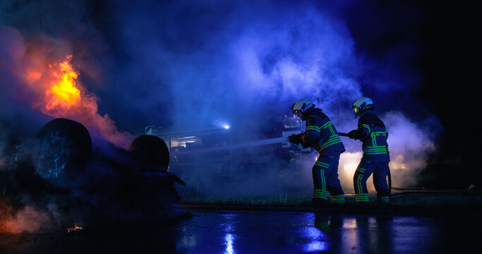 Car Crash. Two Male Firefighters Hold a Hose and Spray Water on Burning Car at Night, Working Next to a Fire Truck to Put Out Flames and Rescue People Trapped After a Serious Traffic Accident.