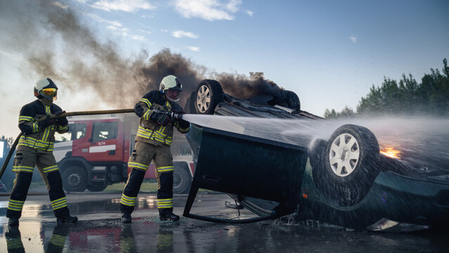 Car Crash. Two Male Firefighters Spray Water on a Burning Overturned Car After a Traffic Collision, Working Beside a Fire Truck to Extinguish Flames and Reach Victims Trapped in the Crashed Vehicle.
