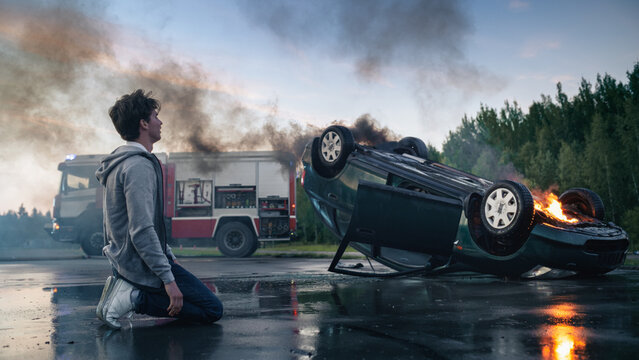 Young Man in Hoodie Kneels on the Road Looking Away From a Burning Overturned Car After a Traffic Accident, Fire Truck With Rescue Workers Arrives at the Car Crash Scene in the Evening