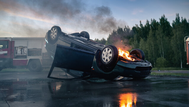 Overturned Burning Car Engulfed in Flames and Smoke on Road After Traffic Accident, Fire Truck With Rescue Workers Arriving at Car Crash Scene to Extinguish Vehicle Fire at Dusk