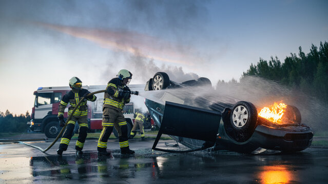 Two Firefighters Spray Water From Hose to Put Out Flames on Rolled Over Car After Traffic Accident, Rescue Workers and Fire Truck at Car Crash Scene on Highway During Emergency Response at Dusk