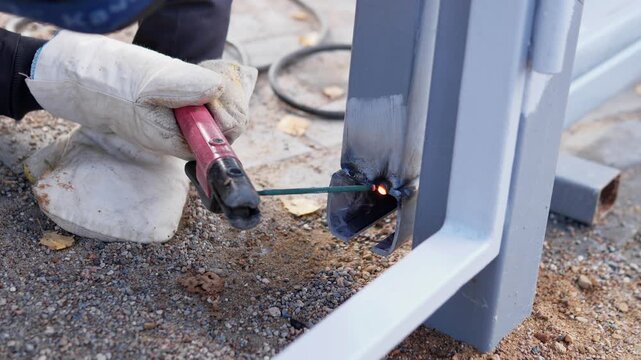 Close-up outdoor repair scene shows welding contact on gray metal post, glowing seam, heat marks, and tiny sparks over rough gravel, with welding, welder, glove, hand detail, showing holder