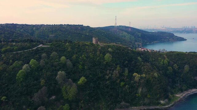 Cinematic drone approach from the Bosphorus strait, flying over the ancient stone ruins of Yoros Castle perched on a lush green hill during spring, with the coastal village of Anadolu Kavagi below