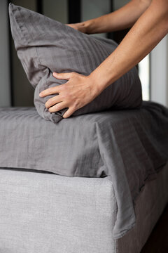Hands fitting a grey sheet on a white mattress. Zamioculcas plant near the bed. Made bed with a blanket and soft pillow visible nearby. Home routine, bedroom tidying set for good sleep. 