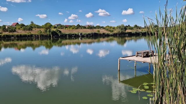 Picturesque lake under blue sky with clouds, place for fishing, country vacation, unity with nature. Landscape with reflection of clouds in water, reeds and village houses on the far shore