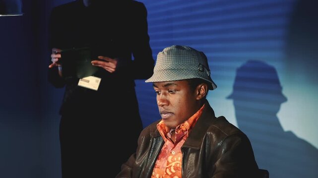 An accused black man wearing a fedora and leather jacket covers his face with hands during an interrogation in a room with a patterned backdrop and standing officer.
