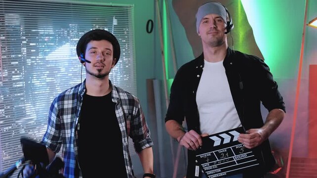 Two adult males preparing for a film shoot indoors, one holding a professional camera and the other a clapper board, with studio lighting and city view behind them.