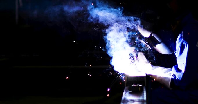 Industrial technician engineer in blue protective cloth wear safety mask, helmet, glasses welding metal. professional welder man welding steel structure with fire spark in factory. selective focus.