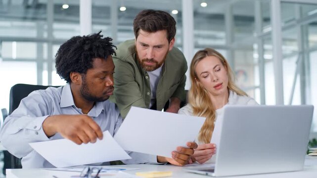 Three multiracial business partners concentrated formal team managing papers documents with laptop corporate service in office colleagues multiethnic group working paperwork analysing using computer
