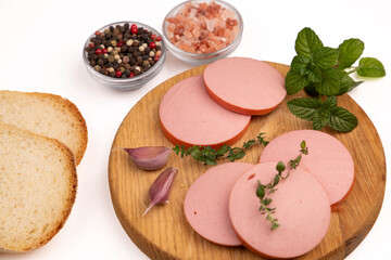 Sliced boiled sausage background closeup with fresh ham slices on wooden cutting board, organic pork meat product with peppercorn mix, pink salt, garlic cloves and mint herbs, white studio e-commerce  © Inna Nikitina