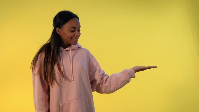 A young woman with long dark hair wearing a light pink hooded sweatshirt smiles and points to the right against a solid yellow studio background.