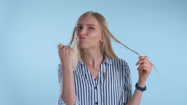 A young blonde woman wearing a striped shirt smiles while playing with her hair in a studio setting with a solid blue background.