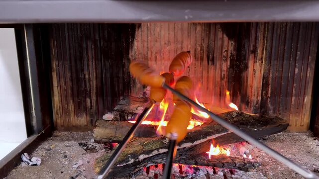 Fire crackles warmly within a rustic fireplace. Sausages sizzle on metal skewers, turning golden over open flame. Embers glow bright beneath the wooden grate. Comforting, cozy atmosphere evokes simple