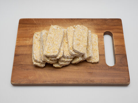 Sliced tempe or tempeh, a traditional Indonesian food made from whole soybeans that are fermented and pressed into a dense, firm cake, on a wooden chopping board with white background