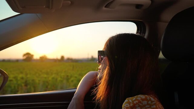 Woman in sunglasses watches farmland through car window at sunset. Female passenger looks at field during road trip. Woman in vehicle viewing countryside. Passenger observes farm at dusk.