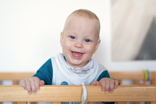 Cheerful baby boy with bright blue eyes smiling joyfully while standing inside a wooden crib during the daytime at home in a cozy nursery room setting