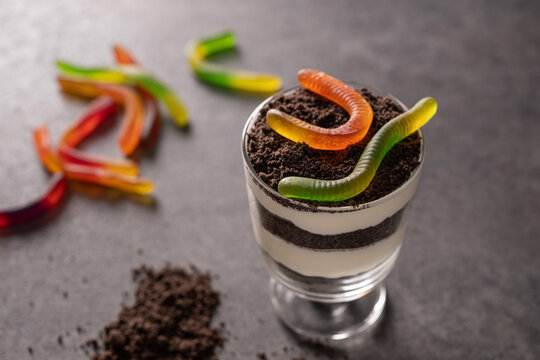 Layered chocolate and cream Dirt cake topped with gummy worms on dark table, styled as a playful spooky dessert for Halloween party.
