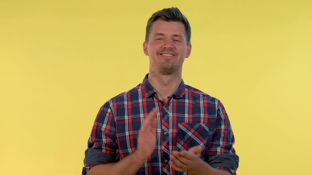 A young man wearing a red, blue, and white plaid shirt smiles and claps his hands against a solid yellow indoor background in a medium close-up shot.