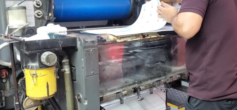 Traditional Offset Printing Press in Action: Quality Control Inspection by Operator.
Fresh offset printed sheets emerge from a traditional printing press as a skilled operator carefully inspects them.