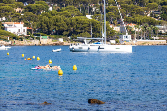 Luxury sailboat anchored in a calm Mediterranean bay while people relax on a paddle board, capturing a peaceful summer vacation and coastal leisure lifestyle