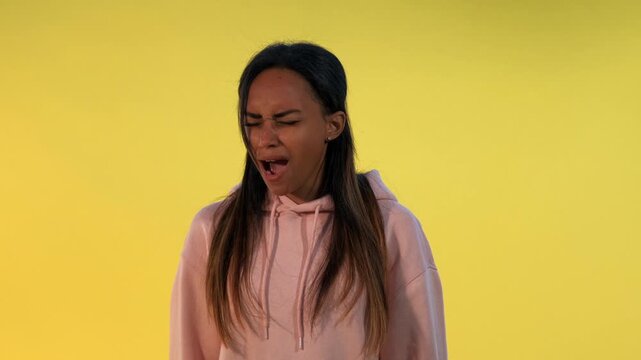 A young woman with long dark hair wearing a light pink hooded sweatshirt is screaming loudly with intense emotion against a solid yellow background in a studio setting.