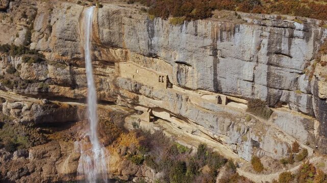 Aerial pullback moving away from the waterfall and cliffside Las Cuevas and San Cornelio hermitages at Yebra de Basa in the Spanish Pyrenees.