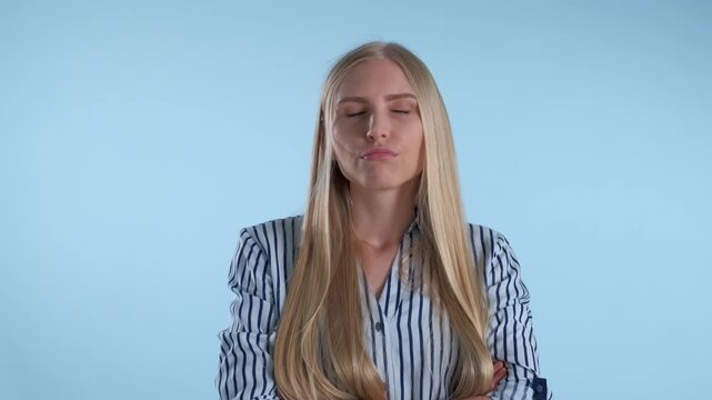 A young blonde woman wearing a striped shirt shows boredom and refusal with crossed arms in a studio with a plain light blue background.