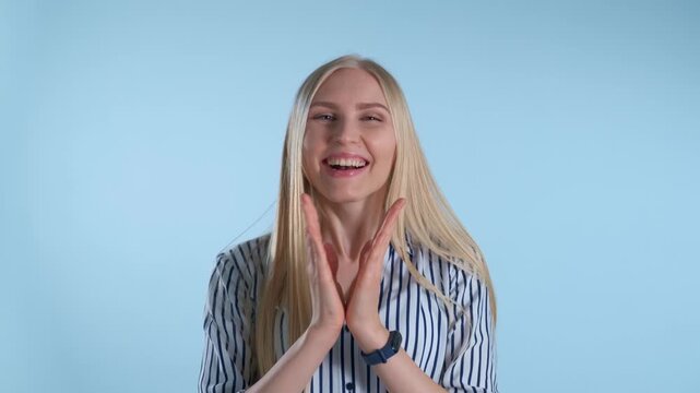 A young blonde woman wearing a blue and white striped shirt claps her hands excitedly while smiling against a solid light blue indoor background.
