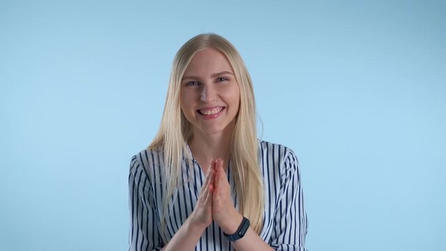 A young blonde woman smiling and clapping her hands indoors against a solid blue background. She wears a striped shirt and a dark wristband on her left wrist.