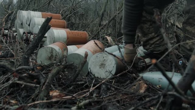 Camouflaged figure walks toward stacked rusty metal canisters in muddy winter woodland, low-angle view with boot steps and branch debris on ground
