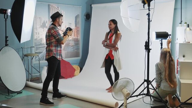 A male photographer directs an adult female model posing in a modern photography studio with assistant and crew amid professional lighting and equipment.