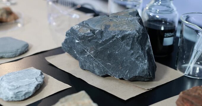 Close-up of rough dark slate rock samples displayed on paper beside glassware and a bottle in a laboratory or geology workspace.