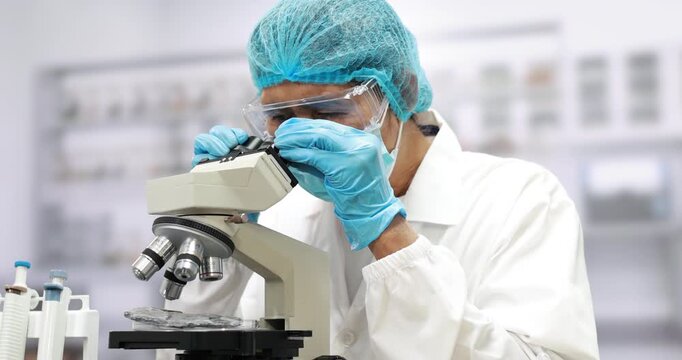 Focused laboratory scientist in protective gloves, mask, goggles and hair cover examining a sample through a microscope in a clean research lab.